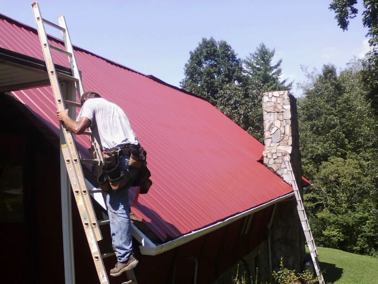 Roofer on a ladder beside a red metal roof and stone chimney — Soffit & Fascia Repair by Roofing Brothers in Kennerdell